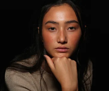 Model with natural makeup, posing with head resting on hand, dark background.