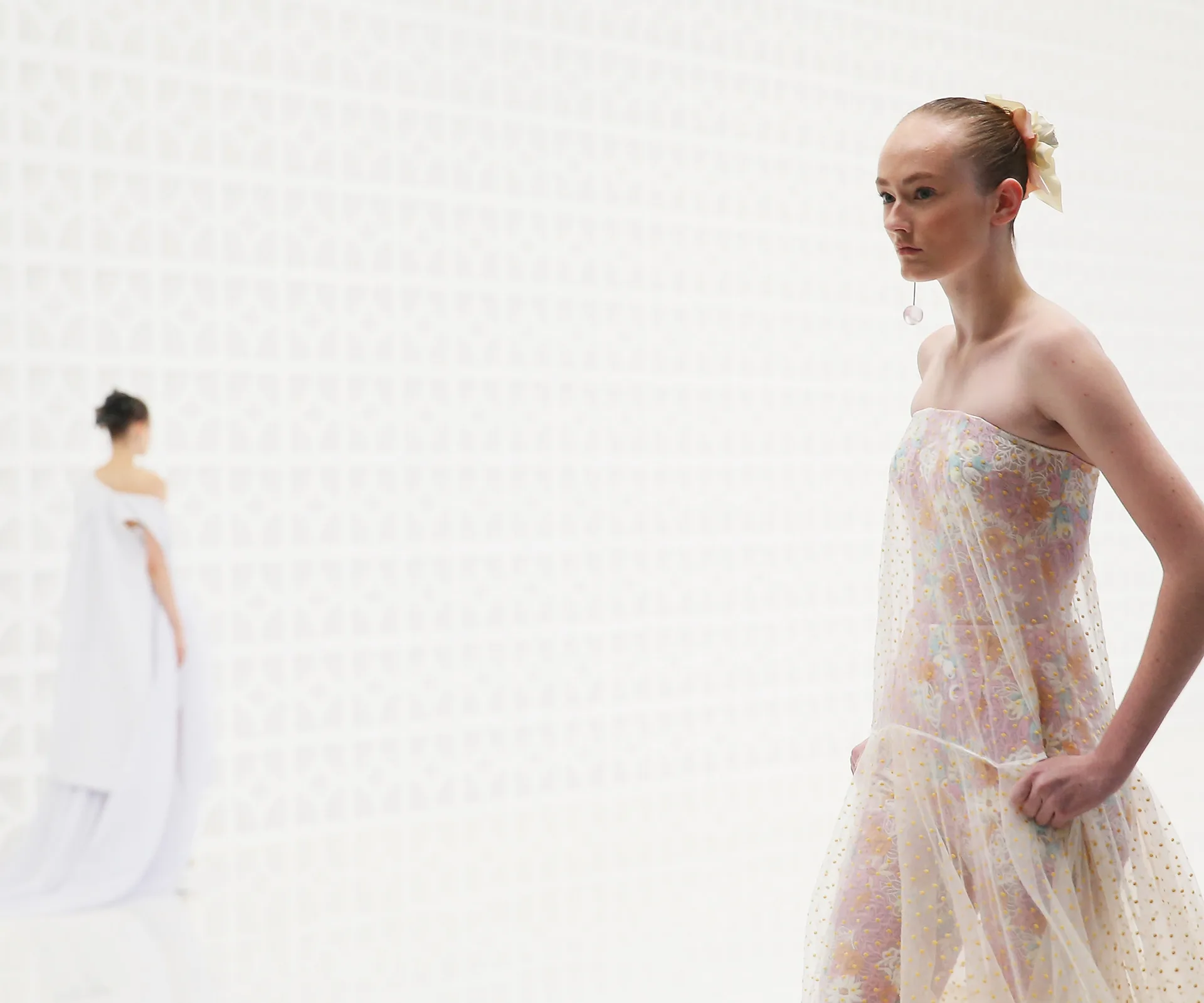 Model in sheer floral dress on runway with geometric patterned backdrop, another model blurred in the background.