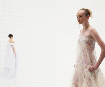 Model in sheer floral dress on runway with geometric patterned backdrop, another model blurred in the background.