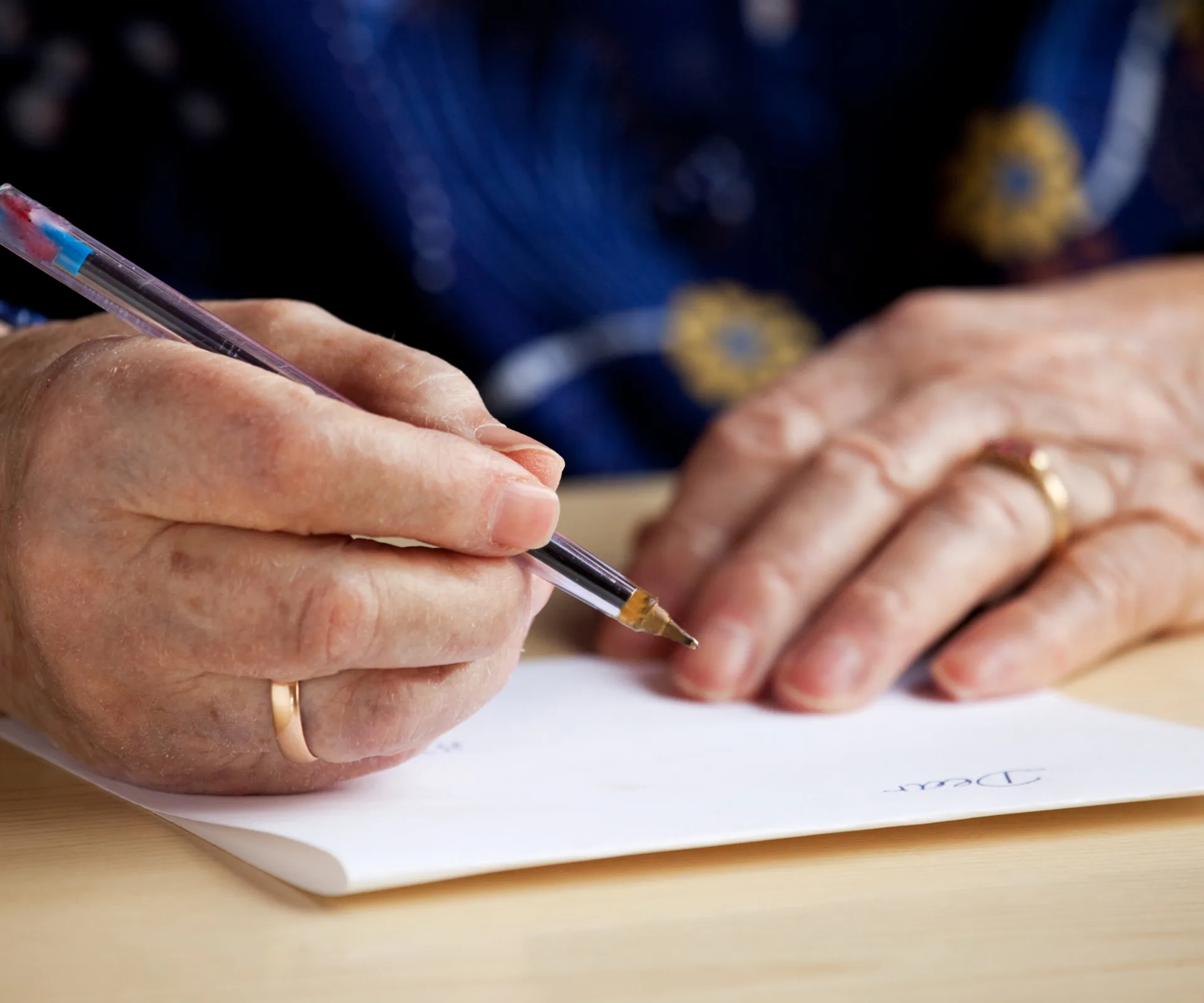 An elderly person writing a letter with a pen on white paper at a wooden table.