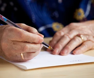 An elderly person writing a letter with a pen on white paper at a wooden table.
