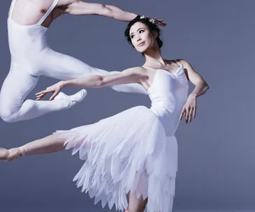 Ballerina in white dress with flowers in hair poses elegantly, while another dancer leaps beside her against a blue backdrop.