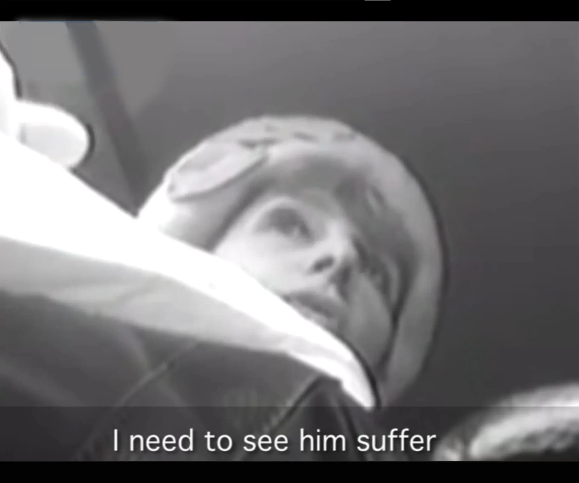 A person in a winter hat captured in a low-angle grayscale surveillance image with subtitles: "I need to see him suffer."