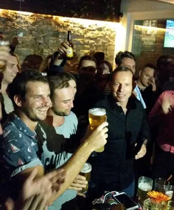 Men at a bar cheers with pints of beer in crowded, lively setting.