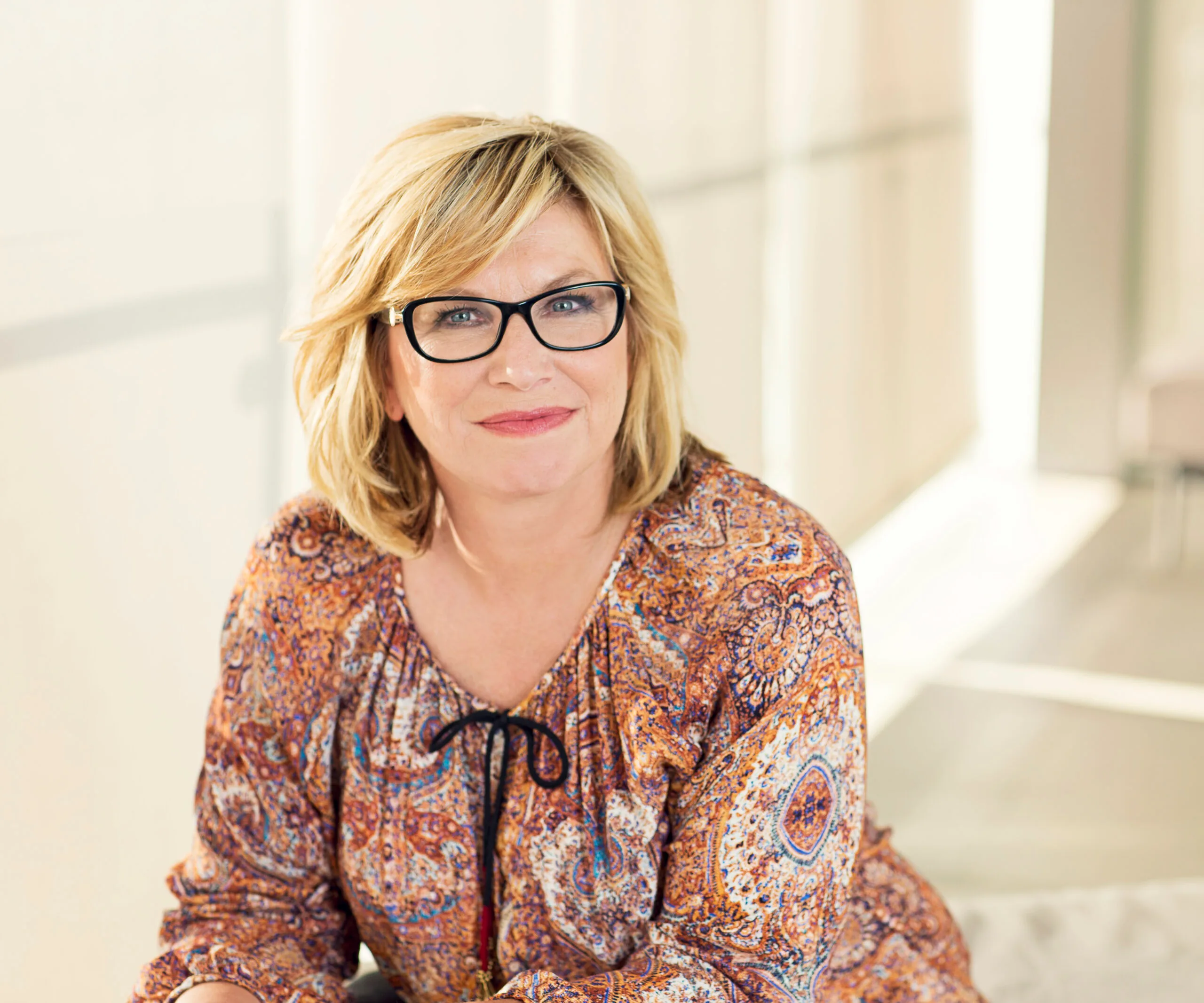 A woman with blonde hair and glasses, wearing a patterned blouse, smiling indoors.