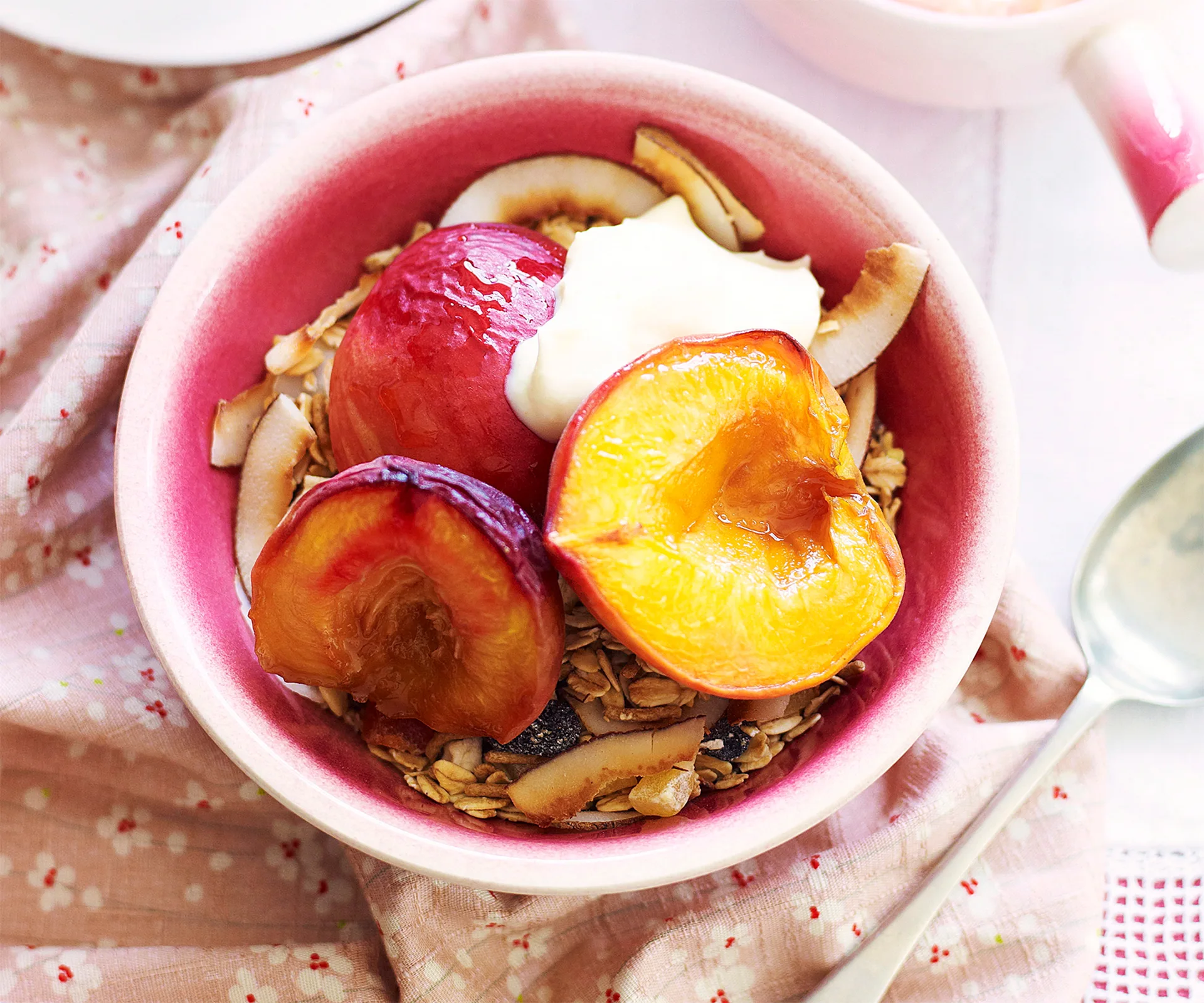 Bowl of granola topped with roasted peaches, coconut flakes, and a dollop of yogurt on a pink cloth.