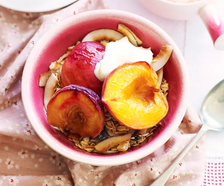 Bowl of granola topped with roasted peaches, coconut flakes, and a dollop of yogurt on a pink cloth.