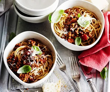 Bowls of spaghetti with lentil Bolognese, topped with basil and parmesan, on a table with forks and a red napkin.