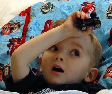 Young boy in a hospital bed holding a toy car, Cars-themed quilt under him, looking upwards in wonder.