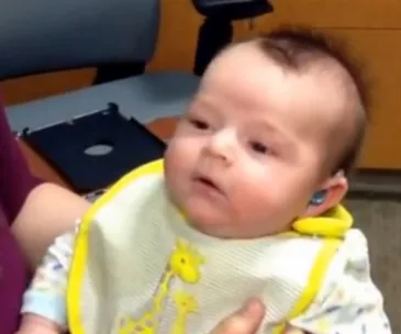 A baby wearing a hearing aid looks surprised while sitting, hearing their mother for the first time.