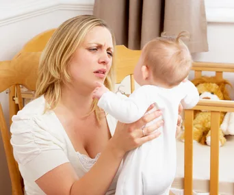 A woman holds an upset baby near a crib, looking concerned. A stuffed lion toy is visible in the background.