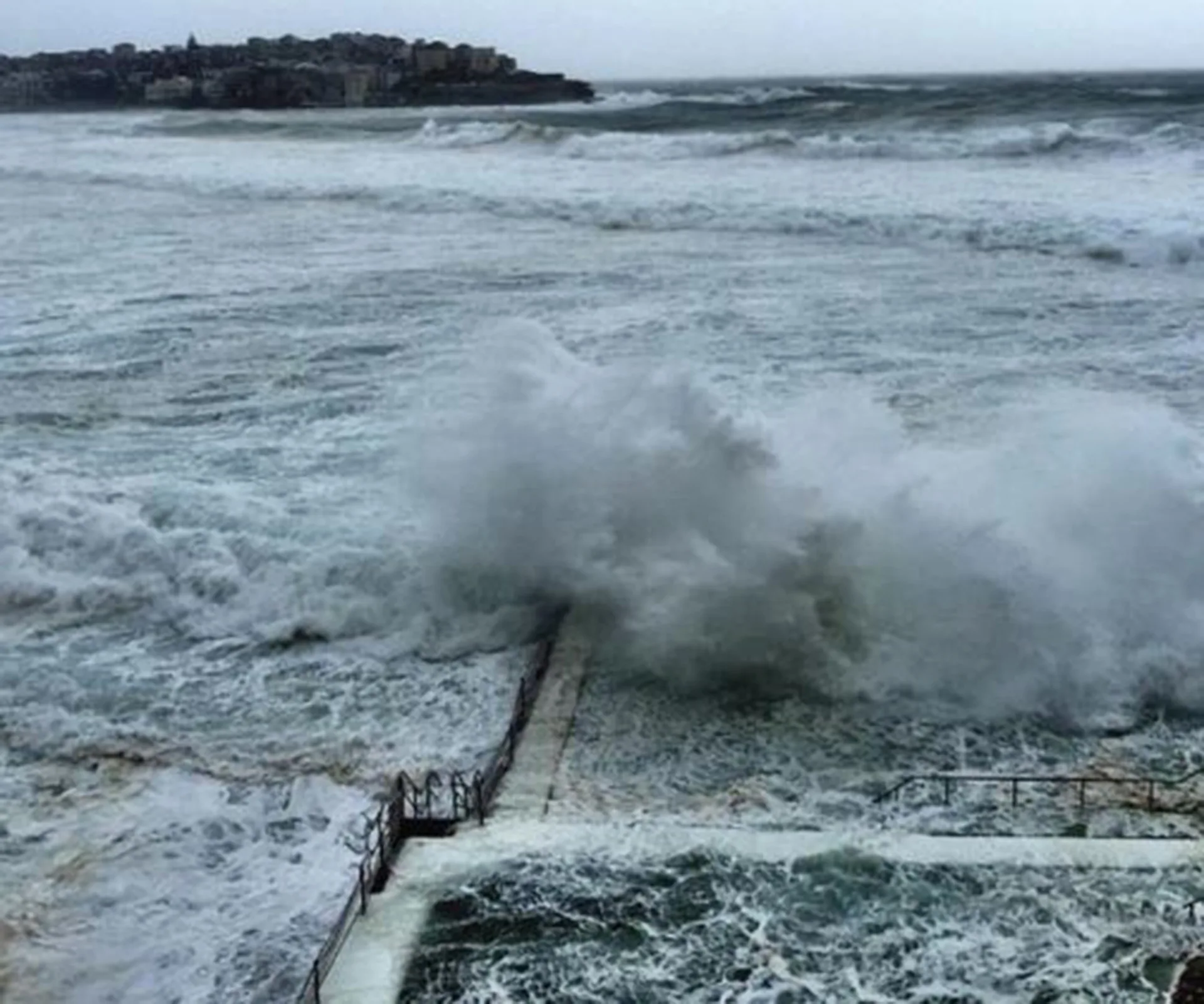 Massive waves crash over Bondi Beach pool during storm, with grey skies and turbulent sea in the background.