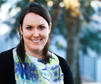 A smiling woman with dark hair in a colorful top, standing outdoors with blurred trees in the background.