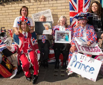 A group of people in Union Jack clothing holding royal-themed signs and photos, celebrating a British royal event.