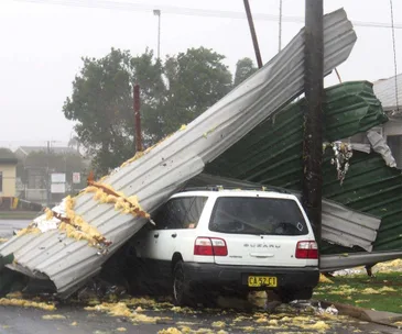 A white Subaru trapped under collapsed metal roof panels on a rainy street, with insulation material scattered around.