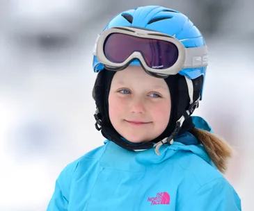 A child in a blue helmet and jacket, wearing ski goggles, smiles against a snowy backdrop.