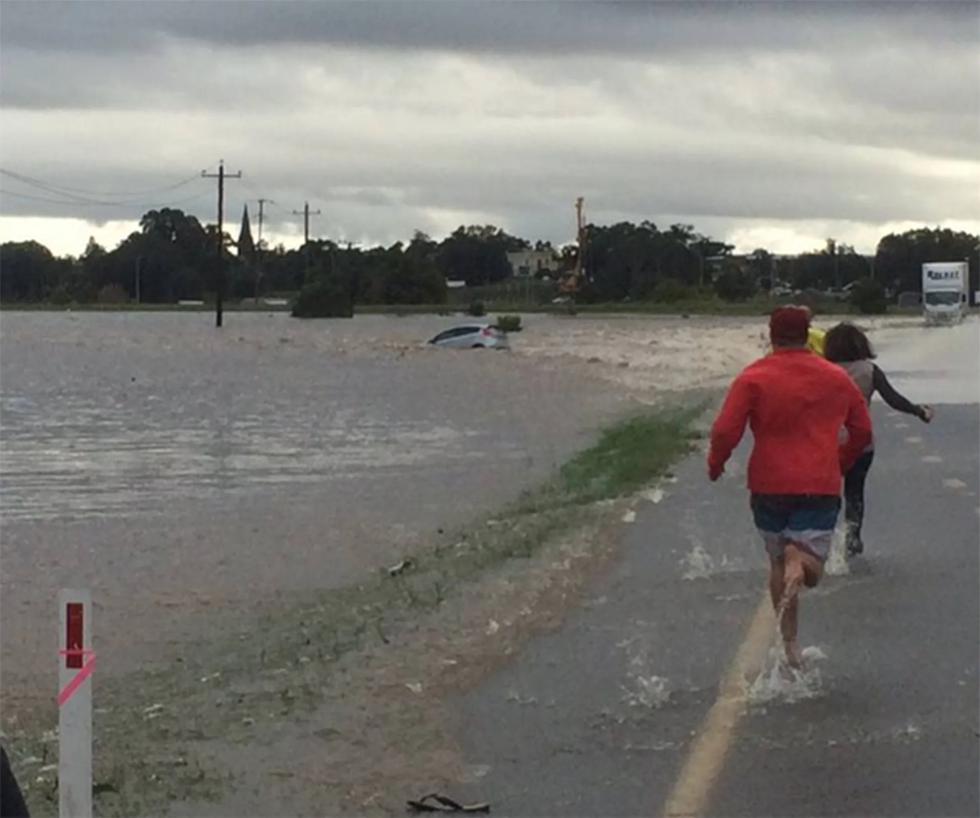 Two people run toward a partially submerged car in a flooded area under a cloudy sky.