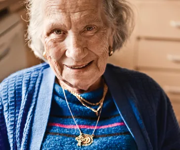 Elderly woman in a blue sweater, smiling warmly, wearing gold necklaces.