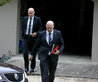Two men in suits walking out of a garage, one holding folders, with a car partially visible in the foreground.