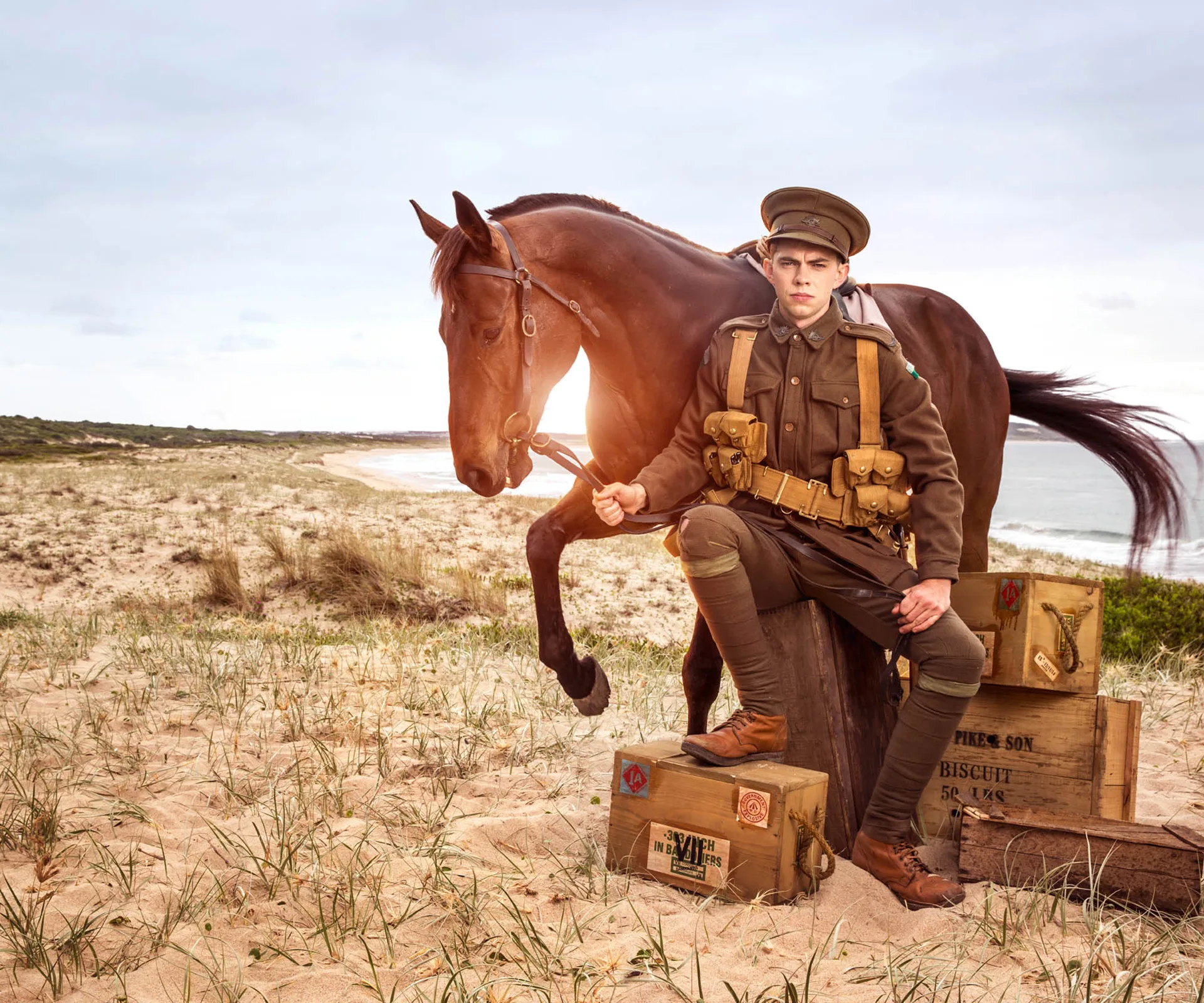 Soldier in WWI uniform with horse on sandy beach, surrounded by wooden crates; symbolizes ANZAC spirit.