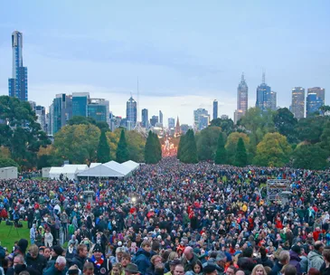 Crowd gathered for Anzac Day ceremony in a park with city skyline in the background, trees lining the path.
