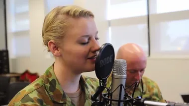 Australian service member in uniform sings into a microphone, another in background playing guitar.