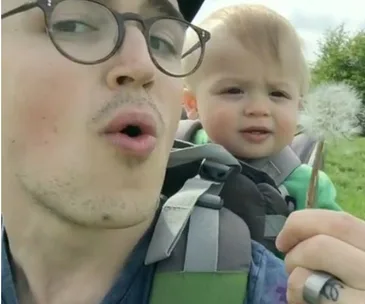 Man blowing on a dandelion with a baby in a carrier on his back, outside in nature.