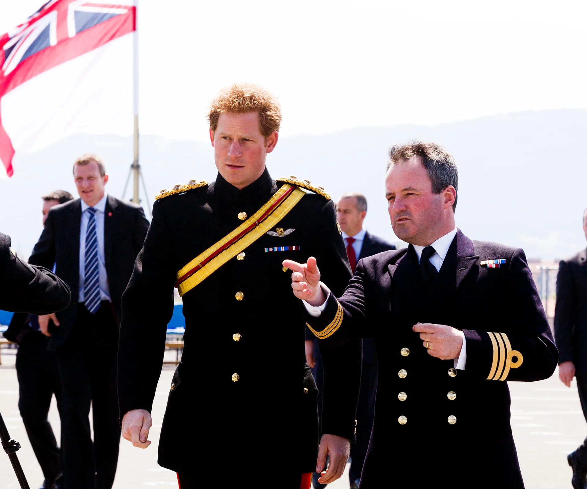 Military personnel in uniform walk alongside British officer, with a flag in the background.