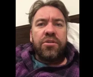 Man with a beard wearing a plaid robe, speaking directly to the camera.