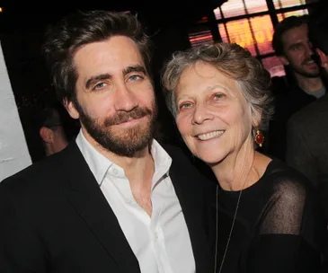 Man with a beard in a suit posing with an older woman wearing earrings, both smiling at an event.