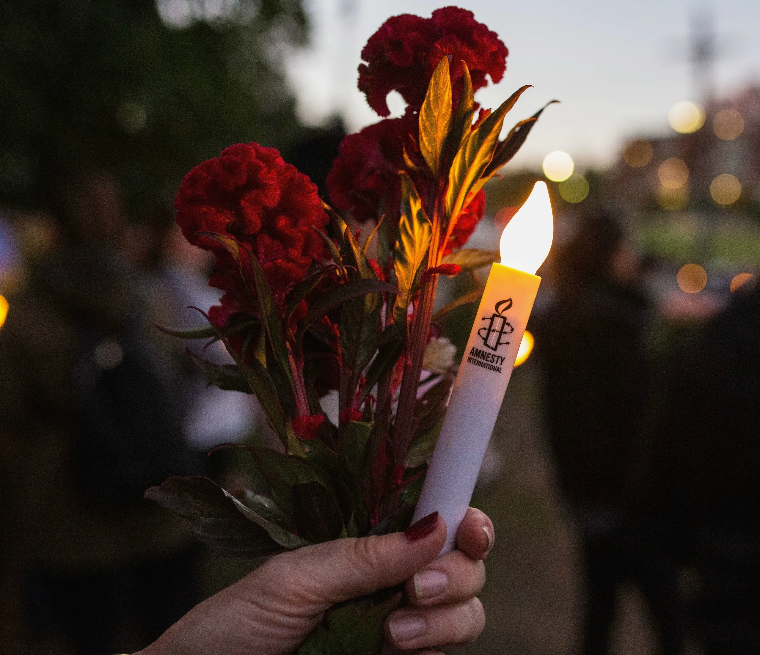 A person holding red flowers and an Amnesty International candle during a vigil at dusk.