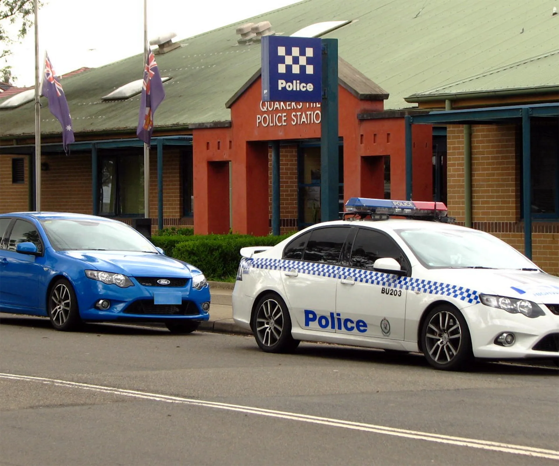 Police station with two parked cars, one police vehicle and one blue sedan, outside a building with Australian flags.