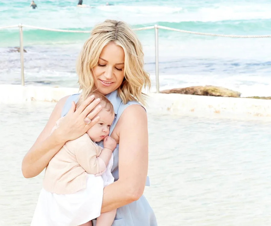Woman holding a baby at the beach, clear ocean waves in the background.