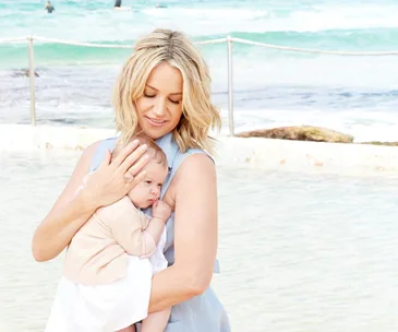 Woman holding a baby at the beach, clear ocean waves in the background.