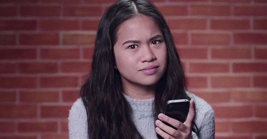 Girl with long hair holding a smartphone, looking puzzled, against a brick wall background.