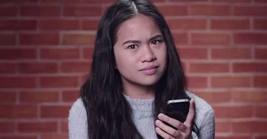 Girl with long hair holding a smartphone, looking puzzled, against a brick wall background.