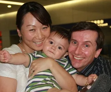 Family smiling, holding a baby wearing a striped shirt, in an indoor setting.