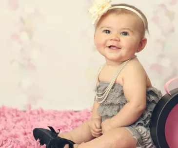 A baby in a lace outfit with pearls and a flower headband sits on a pink rug, wearing small black high heels.