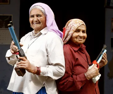 Two elderly women smiling and holding pistols, wearing traditional headscarves and standing back-to-back.