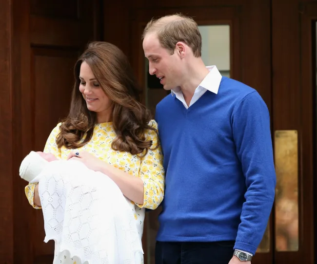 A couple smiling at their newborn wrapped in a white blanket outside a building.