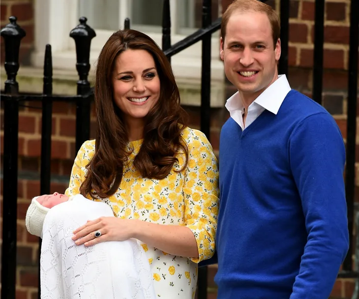 Couple smiling with newborn wrapped in a white blanket outside a building.