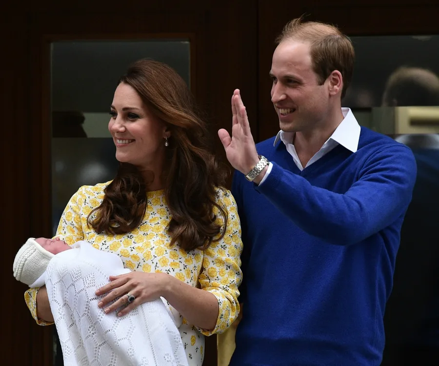 A couple smiling and waving with a newborn wrapped in a white blanket.