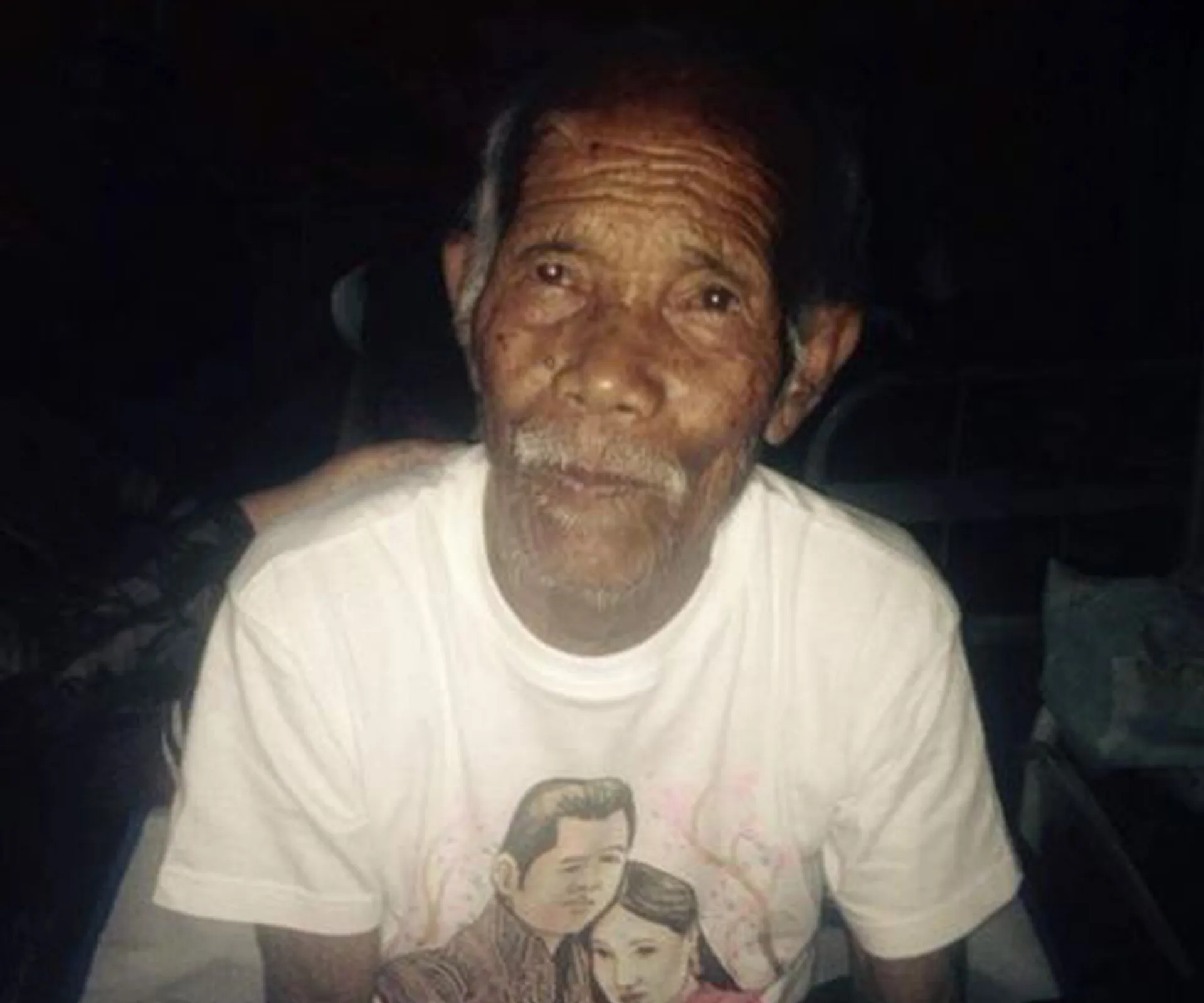 An elderly man with wrinkled skin, wearing a white t-shirt with a graphic print, gazes softly towards the camera.