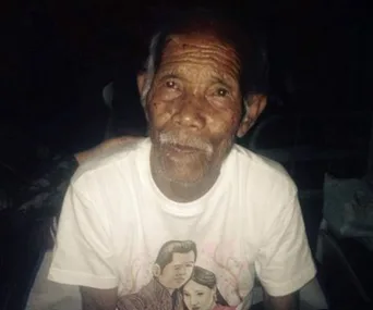 An elderly man with wrinkled skin, wearing a white t-shirt with a graphic print, gazes softly towards the camera.