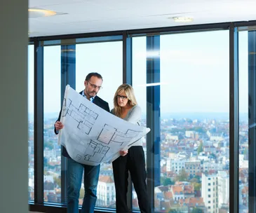 Two people reviewing architectural plans in front of large windows overlooking a cityscape.