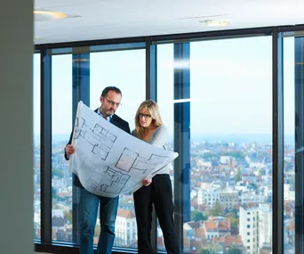 Two people reviewing architectural plans in front of large windows overlooking a cityscape.