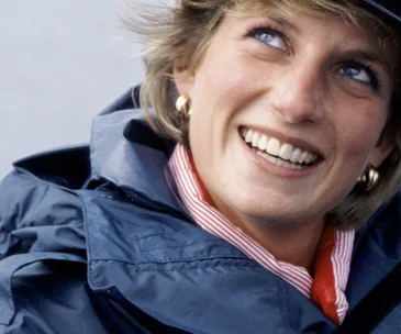A woman smiling warmly, dressed in a navy jacket and red-striped shirt, with short hair and gold earrings.