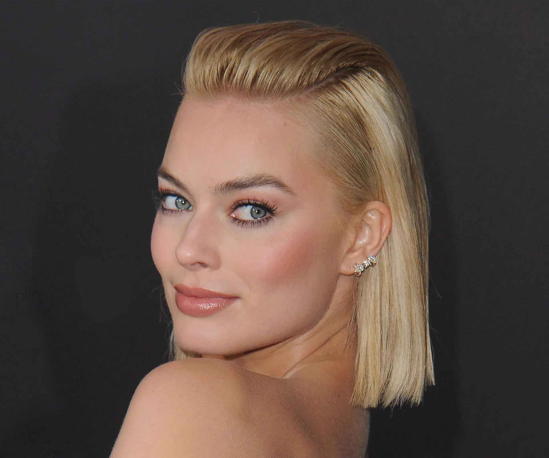 Blonde woman with slicked-back hair, wearing stud earrings, looks over her shoulder against a dark background.