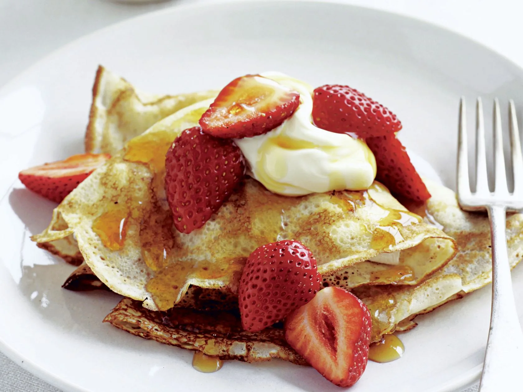 Crepes topped with sliced strawberries, cream, and syrup on a white plate with a fork.