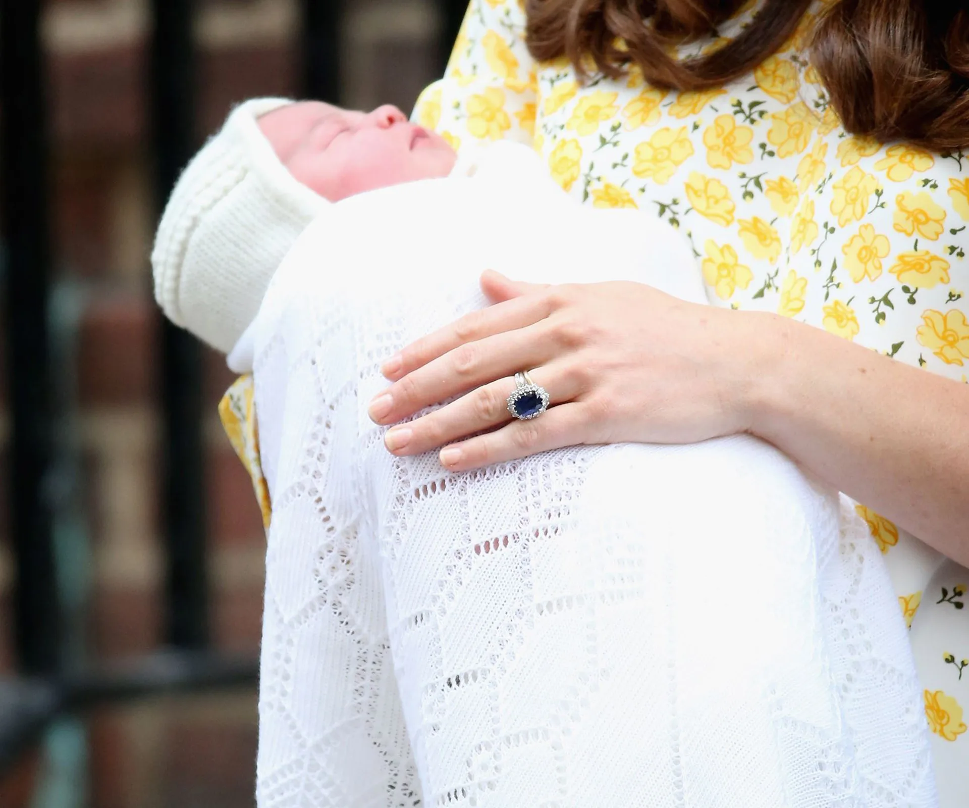 A woman in a floral dress holds a swaddled newborn wearing a knitted hat, hand adorned with a blue sapphire ring.
